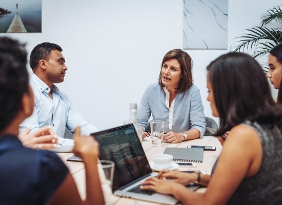 A group of adults sitting around a board table talking and taking notes. 